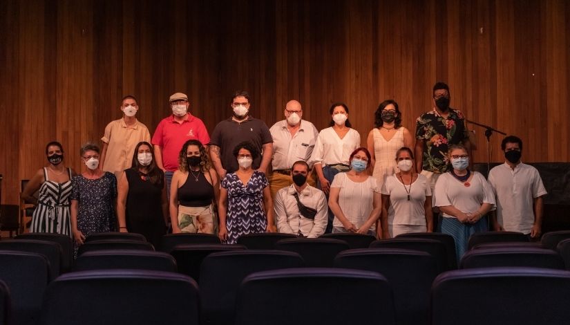 A fotografia mostra os integrantes do CPMLLLB. Estão em pé no palco de um auditório e usam máscaras de proteção respiratória. 